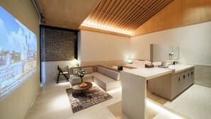Een moderne hotelkamer met een groot projectiescherm, een houten plafond, een bank, een eettafel met een vaas en een keukenhoek.