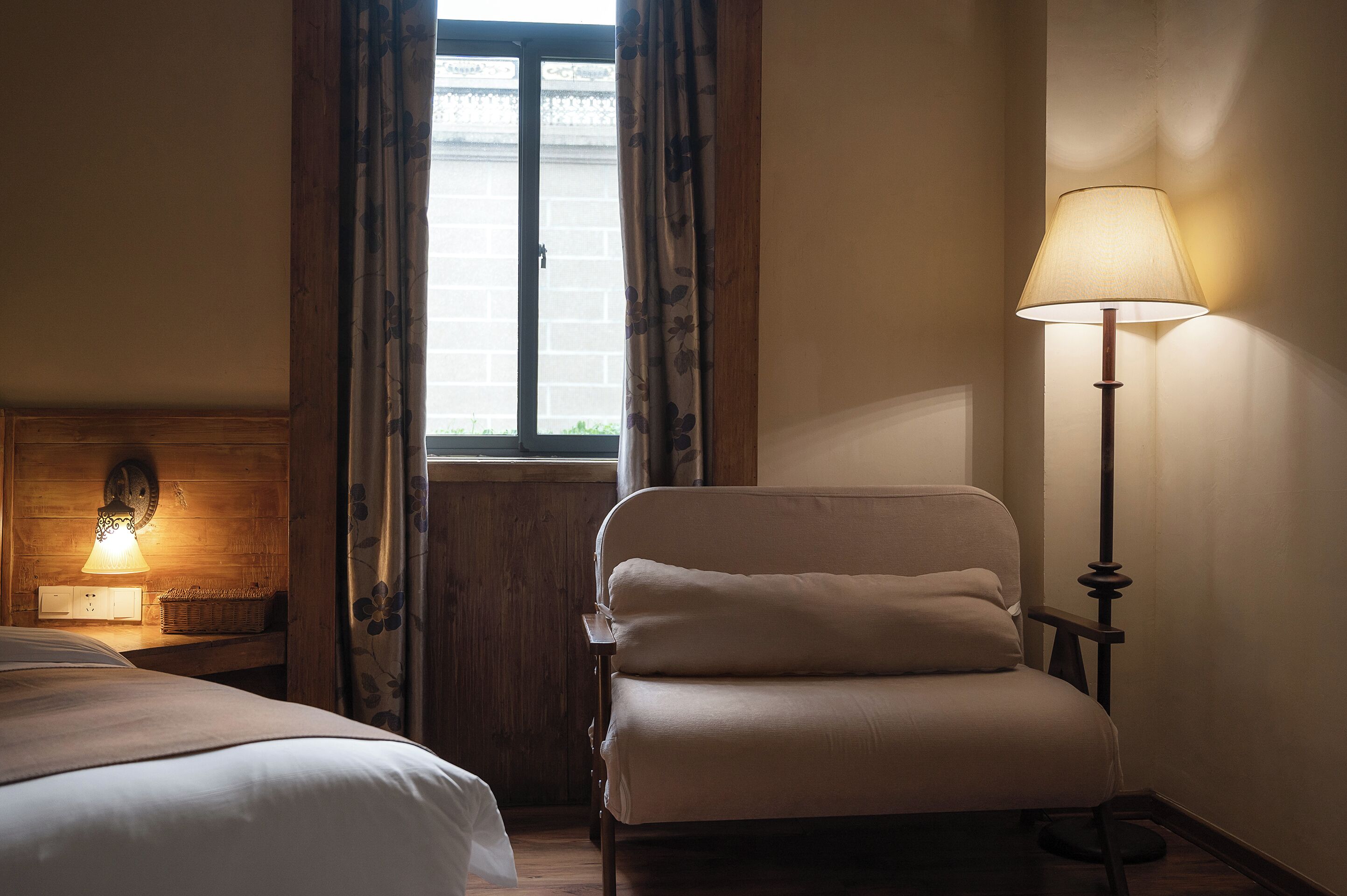 A hotel room with a bed, a chair, a lamp, a window with curtains, and a wall-mounted clock.