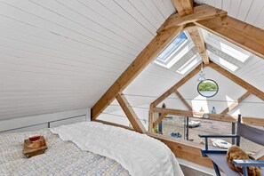A room with a sloped ceiling, a bed with a white blanket, a small table with a bowl, and a view of boats through a window.