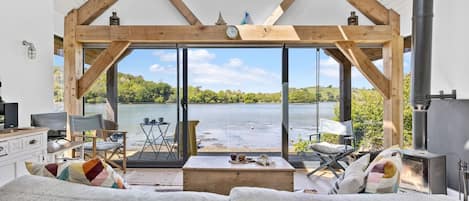 A modern interior with a large window overlooking a body of water, a wooden ceiling structure, and a fireplace.