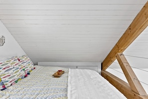 Een slaapkamer met een schuine kap, een bed met beddengoed met een patroon, een nachtkastje met een rode kom, en een houten balkenconstructie.