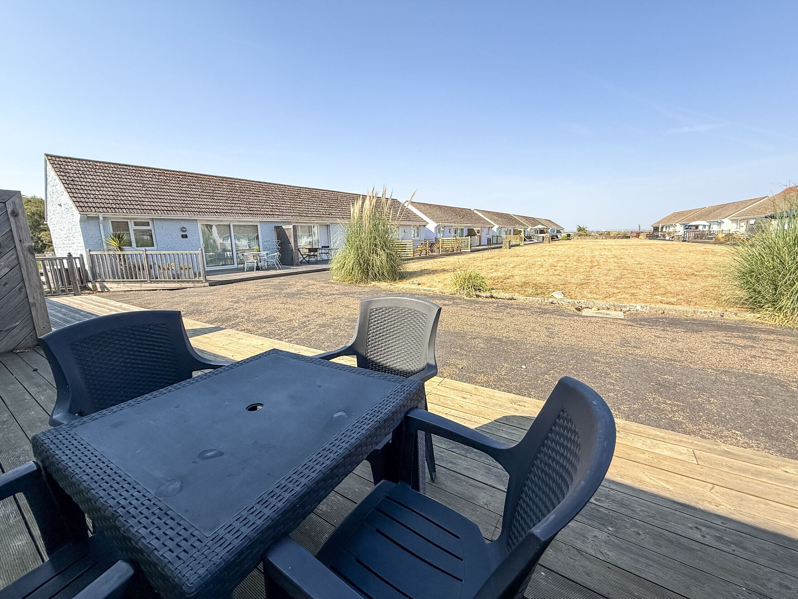 A patio with a table and chairs, a white building with a tiled roof, and a grassy area with houses in the background.