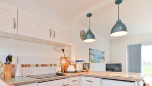 Une cuisine moderne avec des armoires blanches, des comptoirs en granit et deux luminaires suspendus. Un tableau orne le mur et une fenêtre offre une vue sur l’extérieur.
