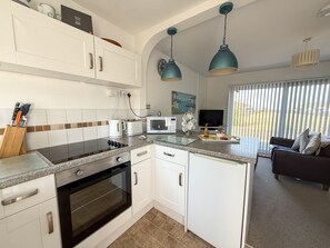 A modern kitchen with white cabinets, a built-in oven, and a dining area with a dark sofa and a TV. - The Hideaway (Seaview)