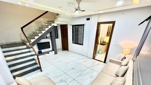 A spacious living area with a marble floor, a sofa set, a staircase, and a kitchen area visible through an open door.