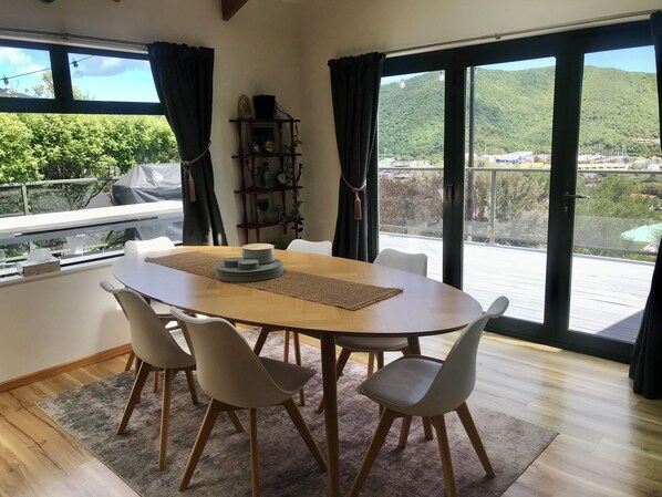 Un coin repas comprenant une table ronde, quatre chaises, une étagère décorative et une vue sur un paysage verdoyant grâce à de grandes fenêtres.