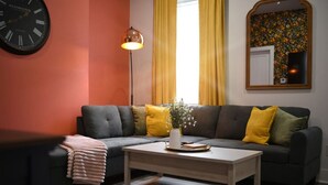 Una sala de estar con un sofá gris oscuro, cojines amarillos, un jarrón blanco con flores, una mesa de centro de madera, un reloj negro, una lámpara de pie y una alfombra estampada.