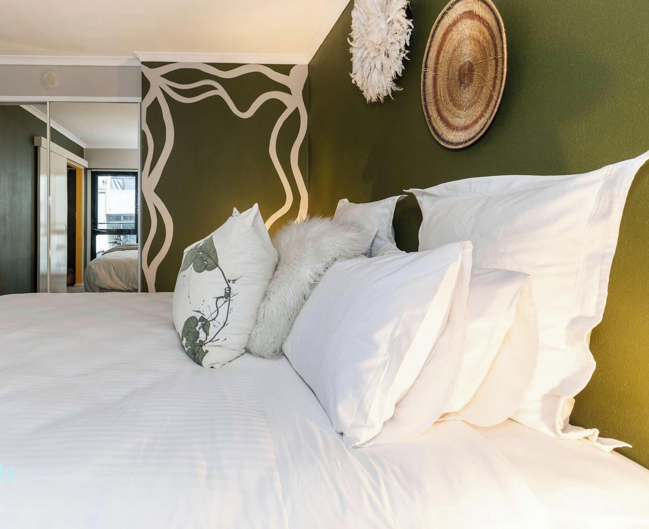 Un lit avec du linge de lit blanc et des coussins décoratifs, une tapisserie murale tissée et une vue sur un balcon.