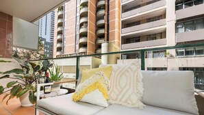 A balcony with a sofa, potted plants, and a building with multiple balconies in the background.