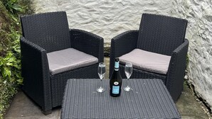Un ensemble comprenant deux fauteuils en osier noir avec des coussins gris et une table carrée assortie, chaque ensemble étant accompagné d'un verre à vin et d'une bouteille de vin.
