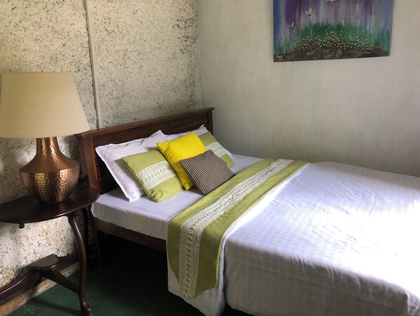 Una cama bien hecha con colcha verde y blanca, un cojín estampado y un cojín amarillo. Hay una lámpara sobre una mesita de noche de madera al lado de la cama.
