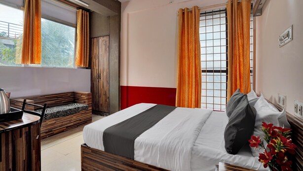 Une chambre d’hôtel avec un lit, un banc en bois, une petite table avec une bouilloire et une fenêtre avec des rideaux orange.