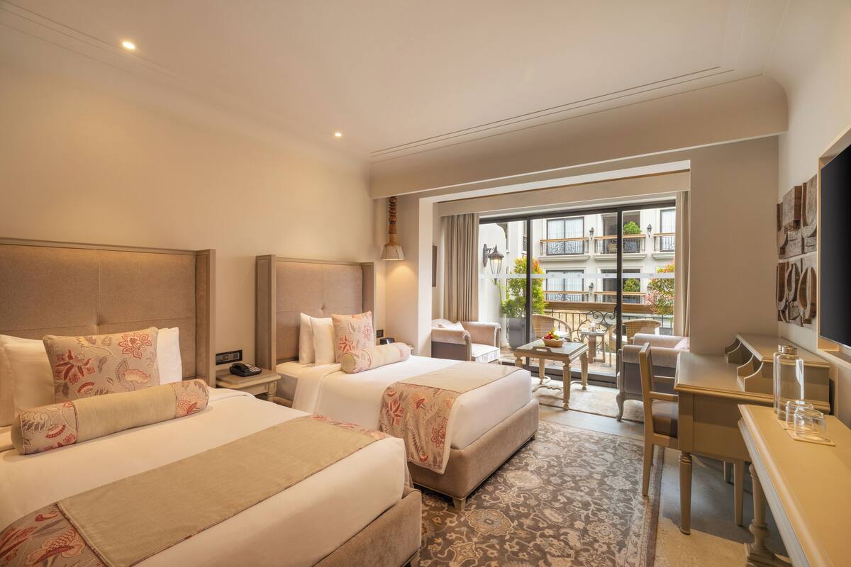 A hotel room with two beds, a desk, and a balcony with a view of the city.