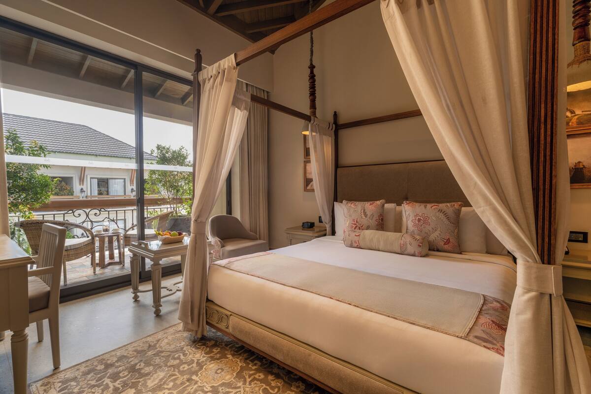 A bedroom with a four-poster bed, a small table with a chair, and a view of the outside.