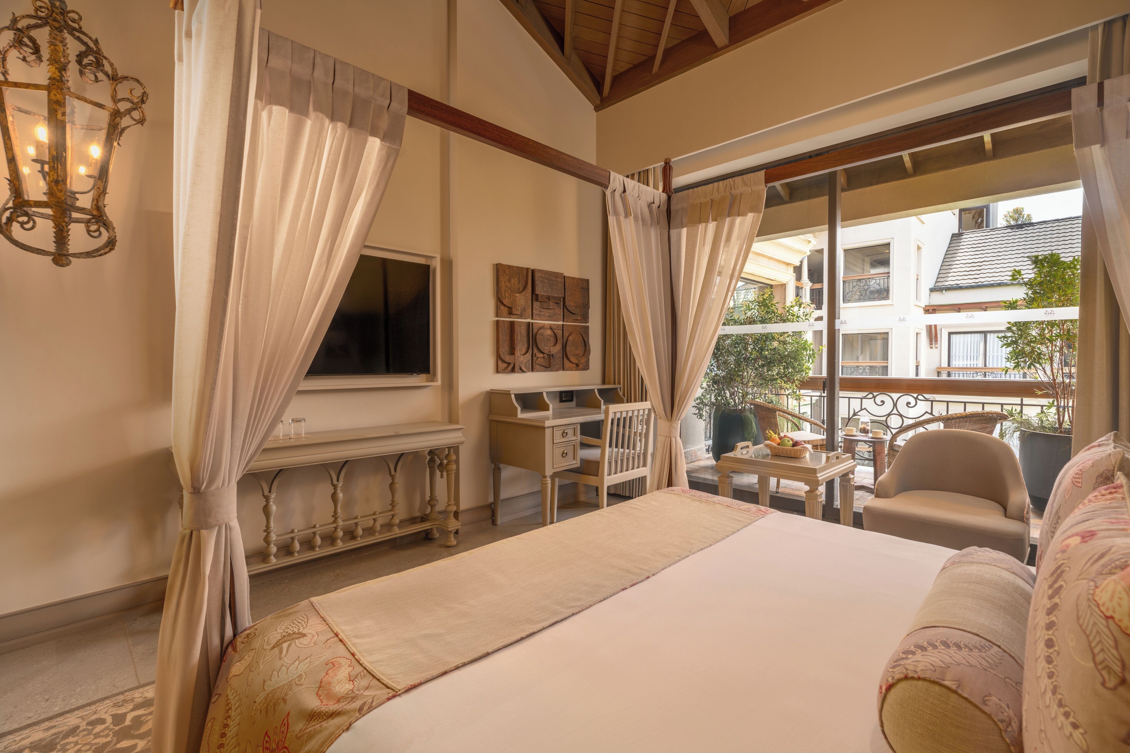 A bedroom with a large bed, a desk, a chair, and a balcony with a view of buildings and greenery.
