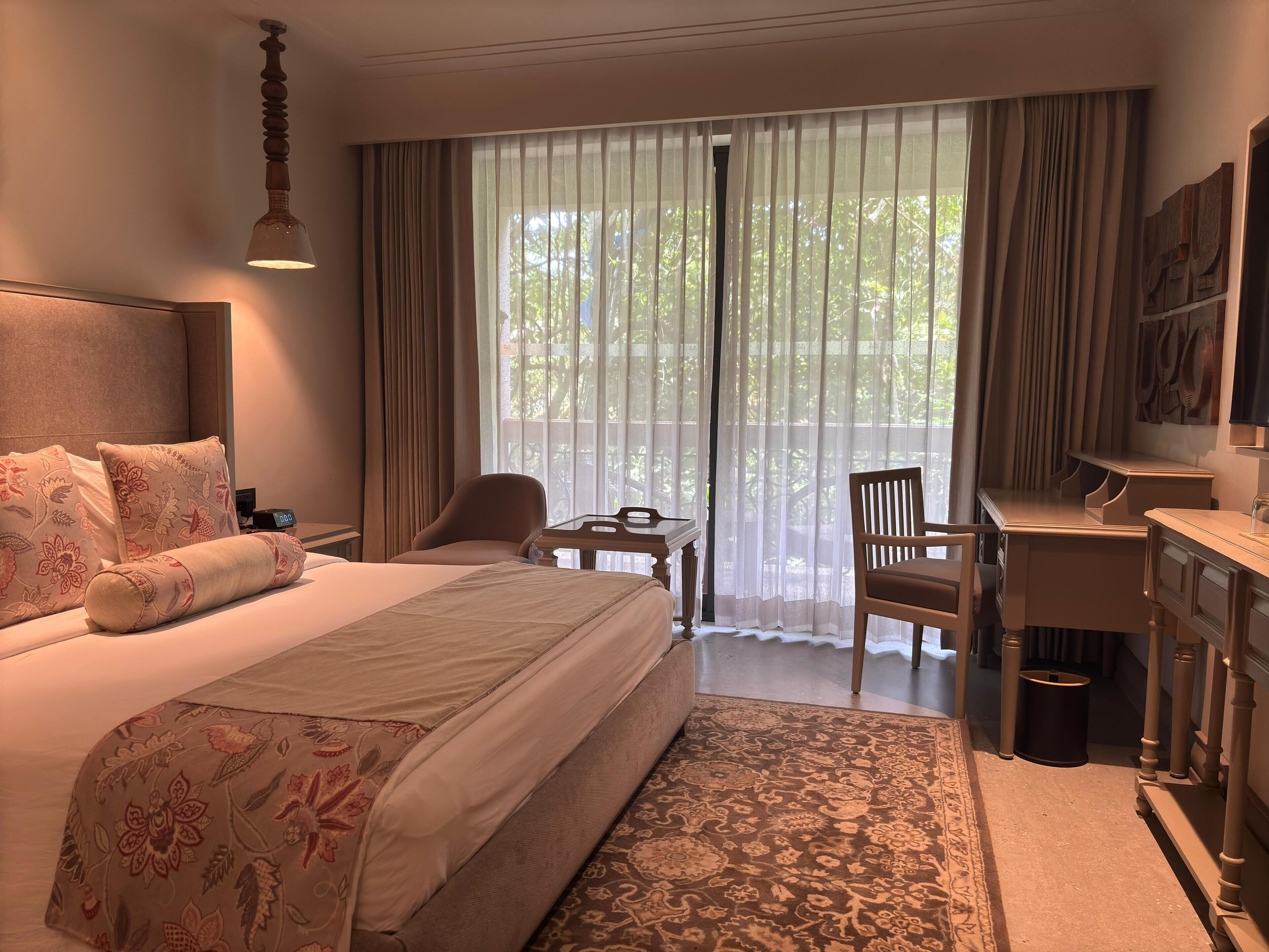 A hotel room with a large bed, a desk, a chair, and a view of greenery through sheer curtains.