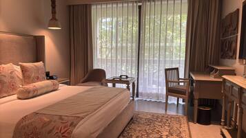 A hotel room with a large bed, a desk, a chair, and a view of greenery through sheer curtains.