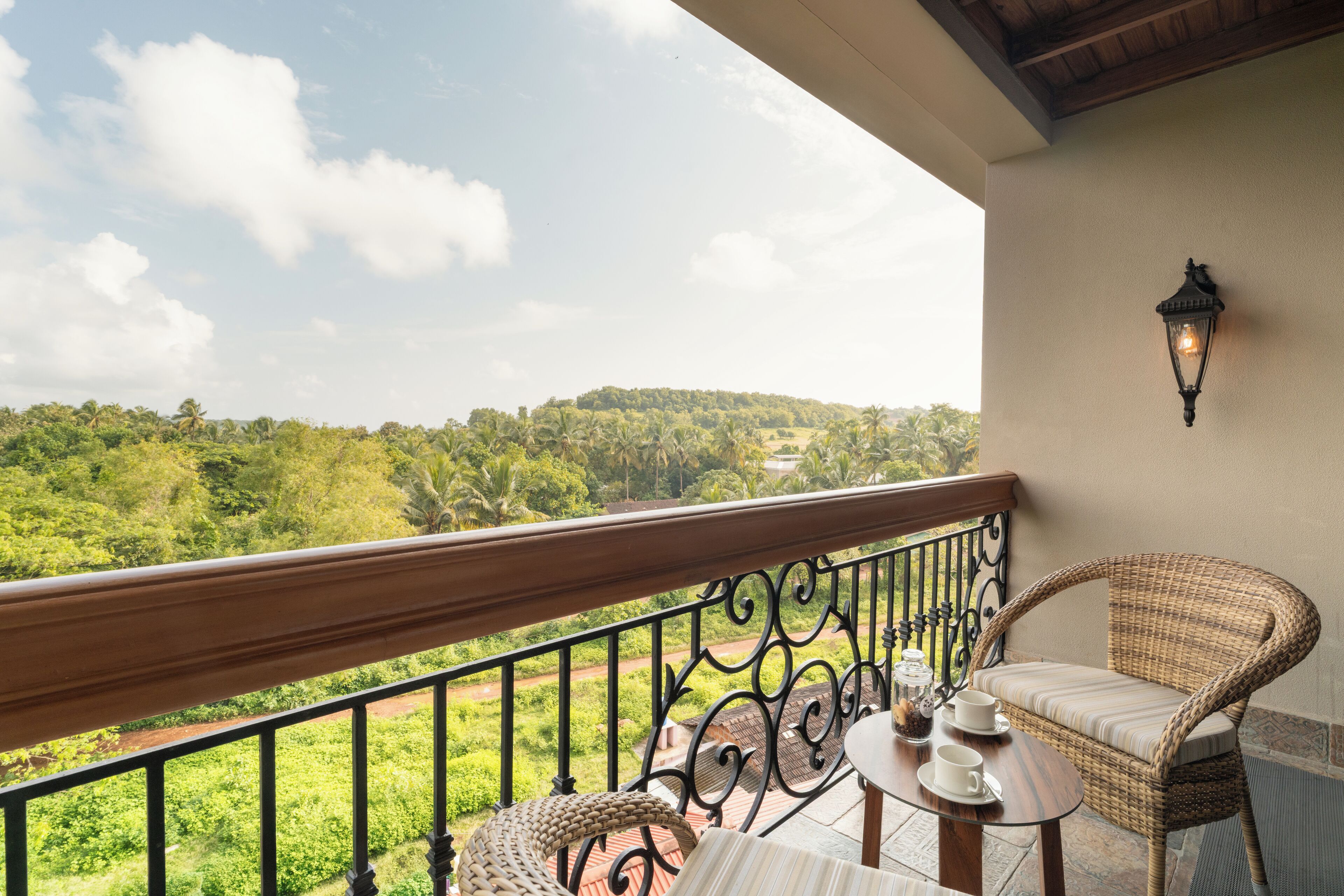 A balcony with a wicker chair, a small table with a cup and saucer, and a view of a green landscape.