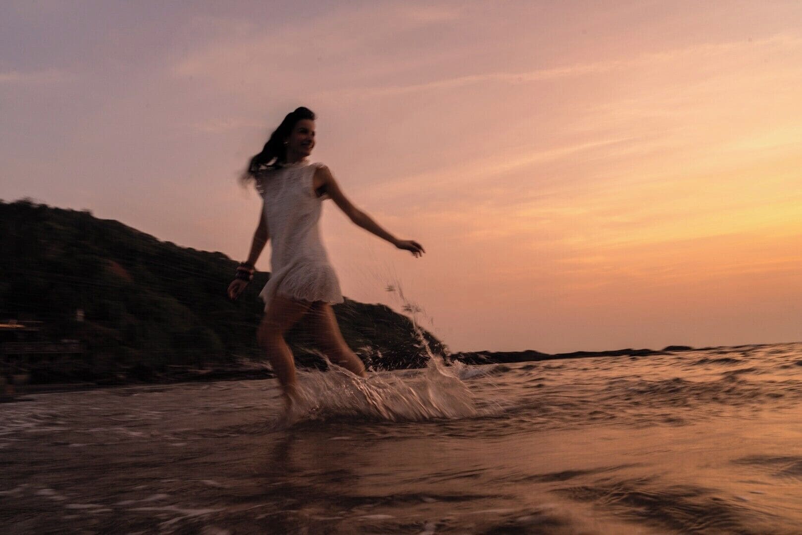 A person in a white dress walking through shallow water near a rocky shoreline during sunset.