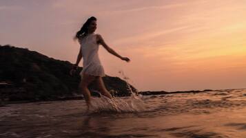 A person in a white dress walking through shallow water near a rocky shoreline during sunset.