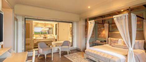 Une chambre à coucher avec un lit à baldaquin, un coin salon avec des fauteuils, un bureau et une vue sur la salle de bain.
