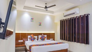 A modern hotel room with a wooden headboard, a bed with colorful pillows, a ceiling fan, an air conditioning unit, and a curtain with a dark drape. - Hotel M K Residency (Pune)