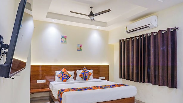 Habitación de hotel moderna con cabecera de madera, cama con almohadas coloridas, ventilador de techo, equipo de aire acondicionado y cortina con un tejido oscuro.