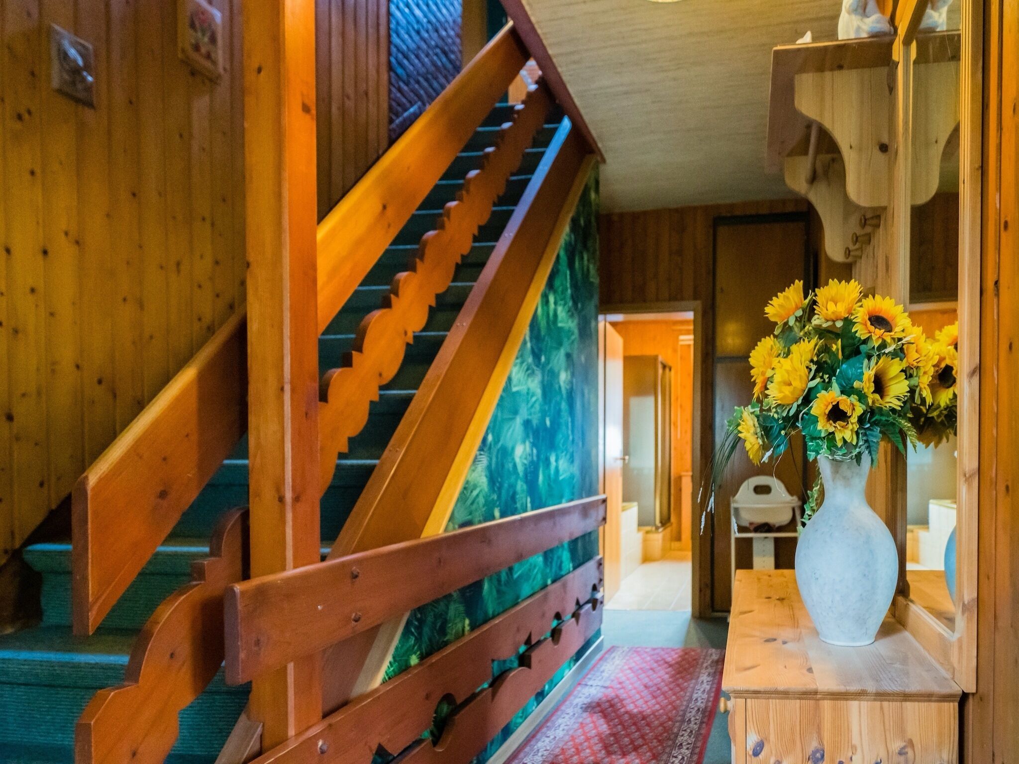 Un escalier en bois avec une rampe, un vase de tournesols posé sur une table en bois et un tapis à motifs sur le sol.
