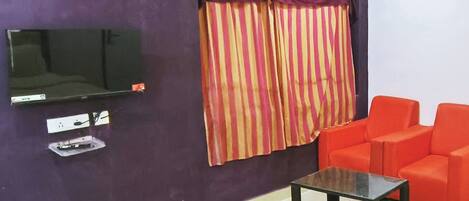 A room with a flat-screen TV mounted on a purple wall, a window with striped curtains, a red sofa, and a small table.
