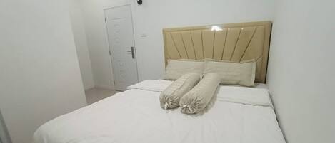 A neatly made bed with a beige headboard and white bedding in a simple, clean room with white walls and a door in the background.