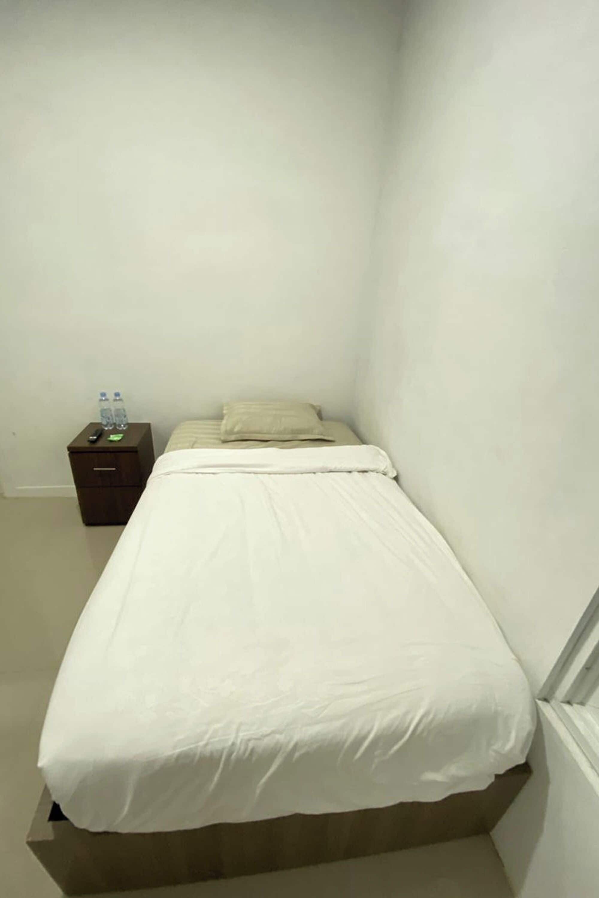 Un petit lit bien fait, avec une couverture blanche, dans une chambre d’angle aux murs blancs.