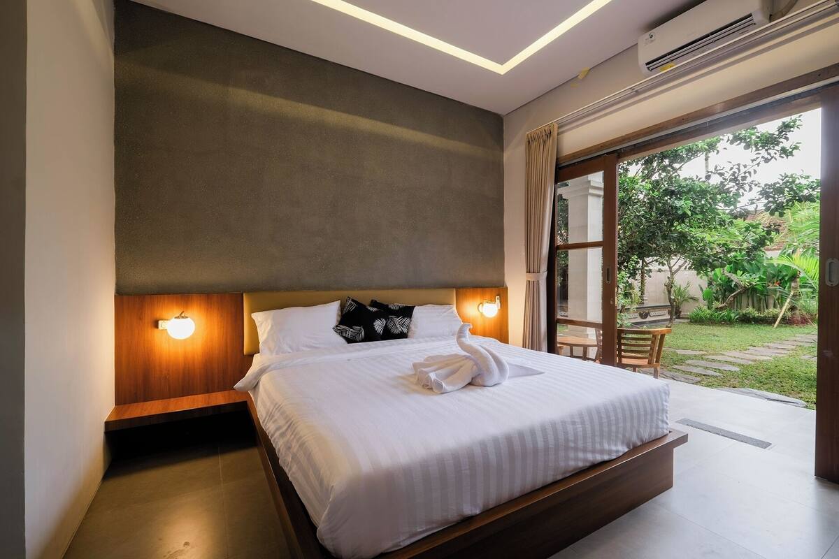 A modern bedroom with a large bed, a wooden headboard, a nightstand with a lamp, and a view of a garden through a sliding door.