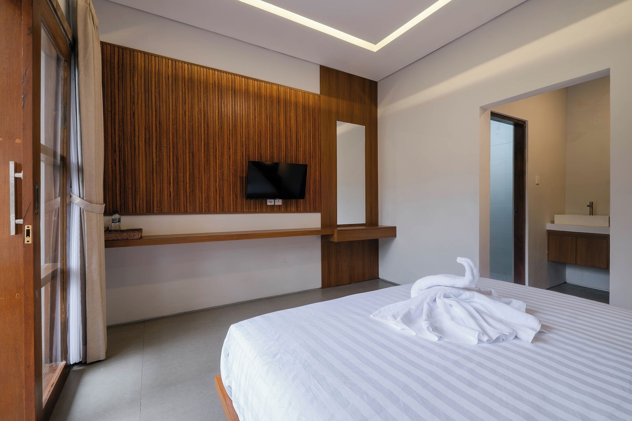 A hotel room with a bed, a TV mounted on a wooden panel wall, a mirror, and a bathroom visible through an open door.