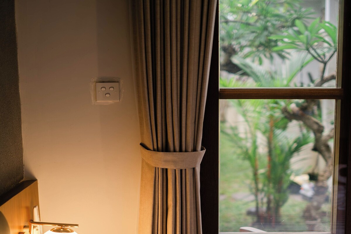 Habitación de hotel con cama, lámpara de noche, ventana con cortinas y vistas al exterior.