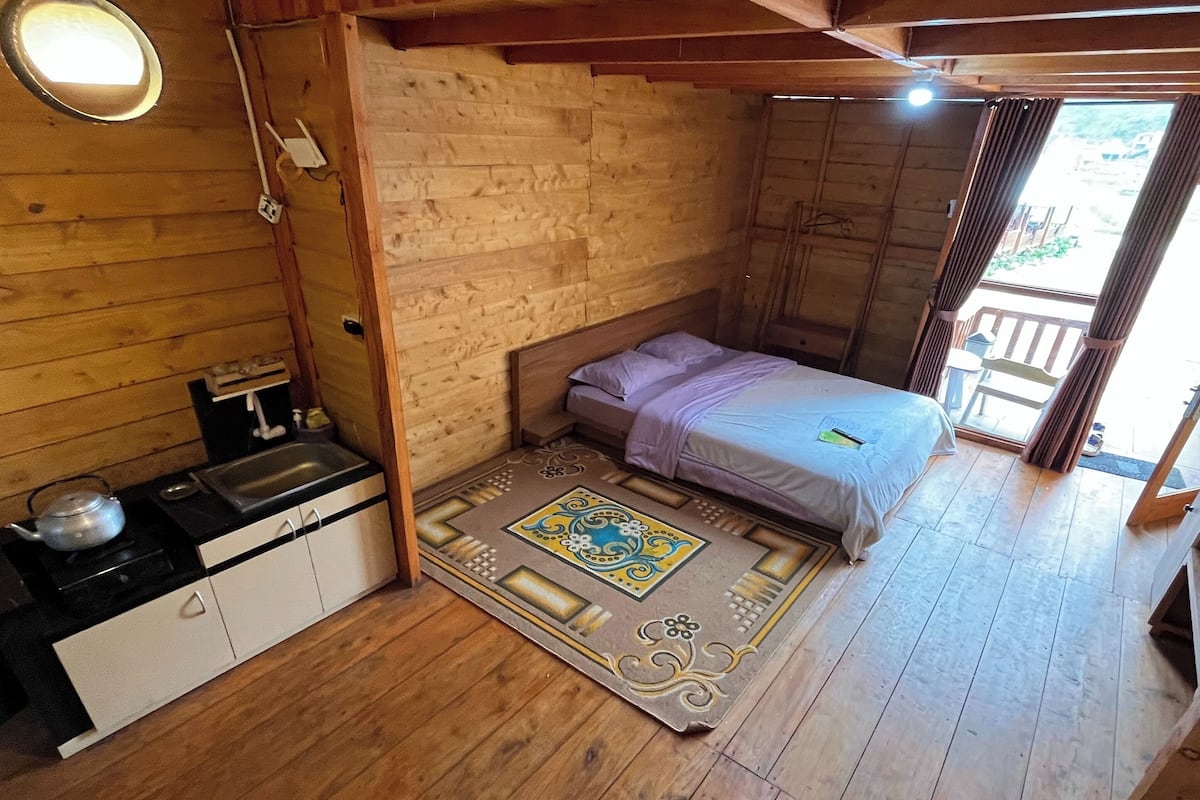 A small, cozy room with a wooden ceiling, a bed with a purple blanket, a small kitchenette, and a window with curtains.