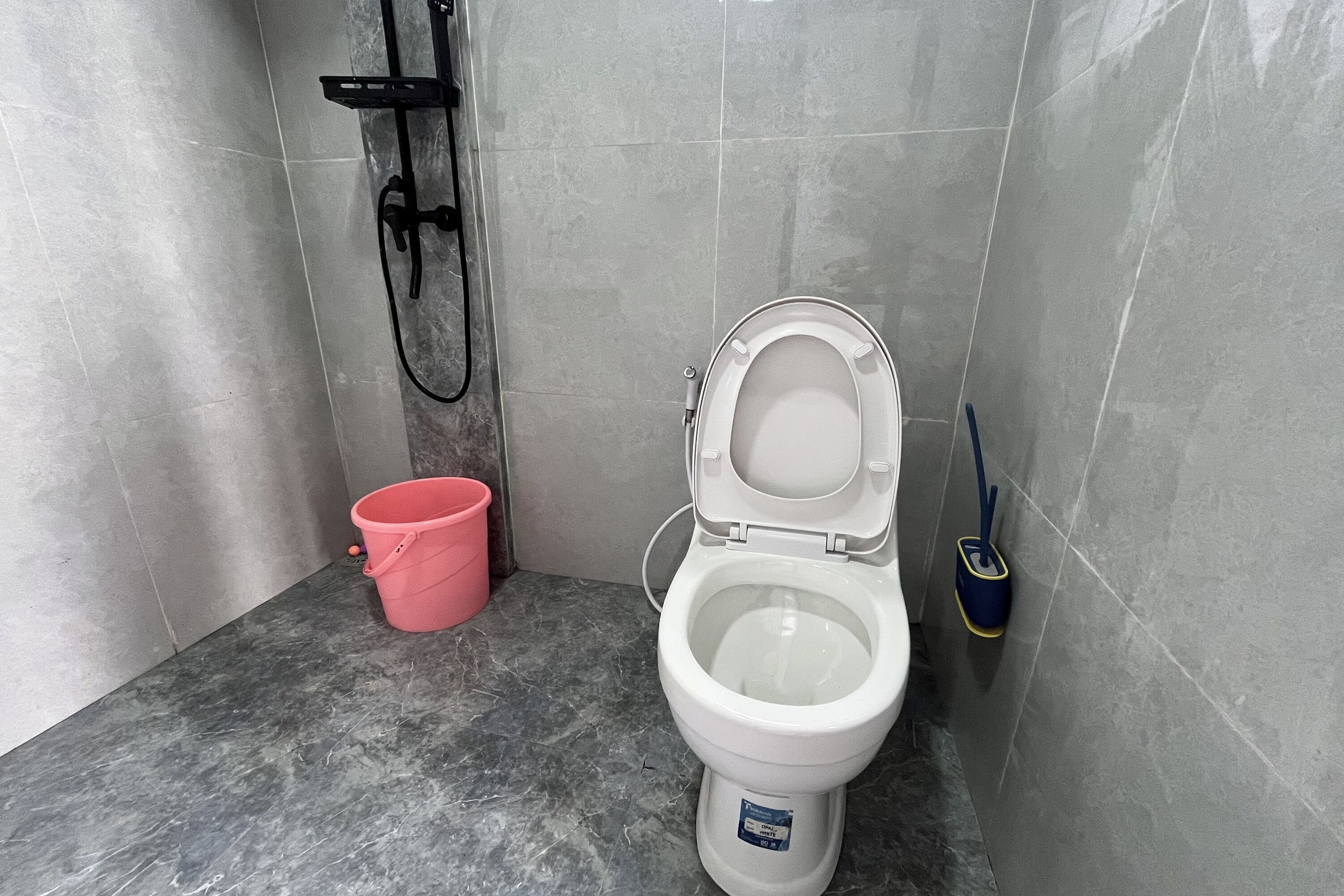 Casa de banho com sanita branca, balde rosa e escova de limpeza azul.