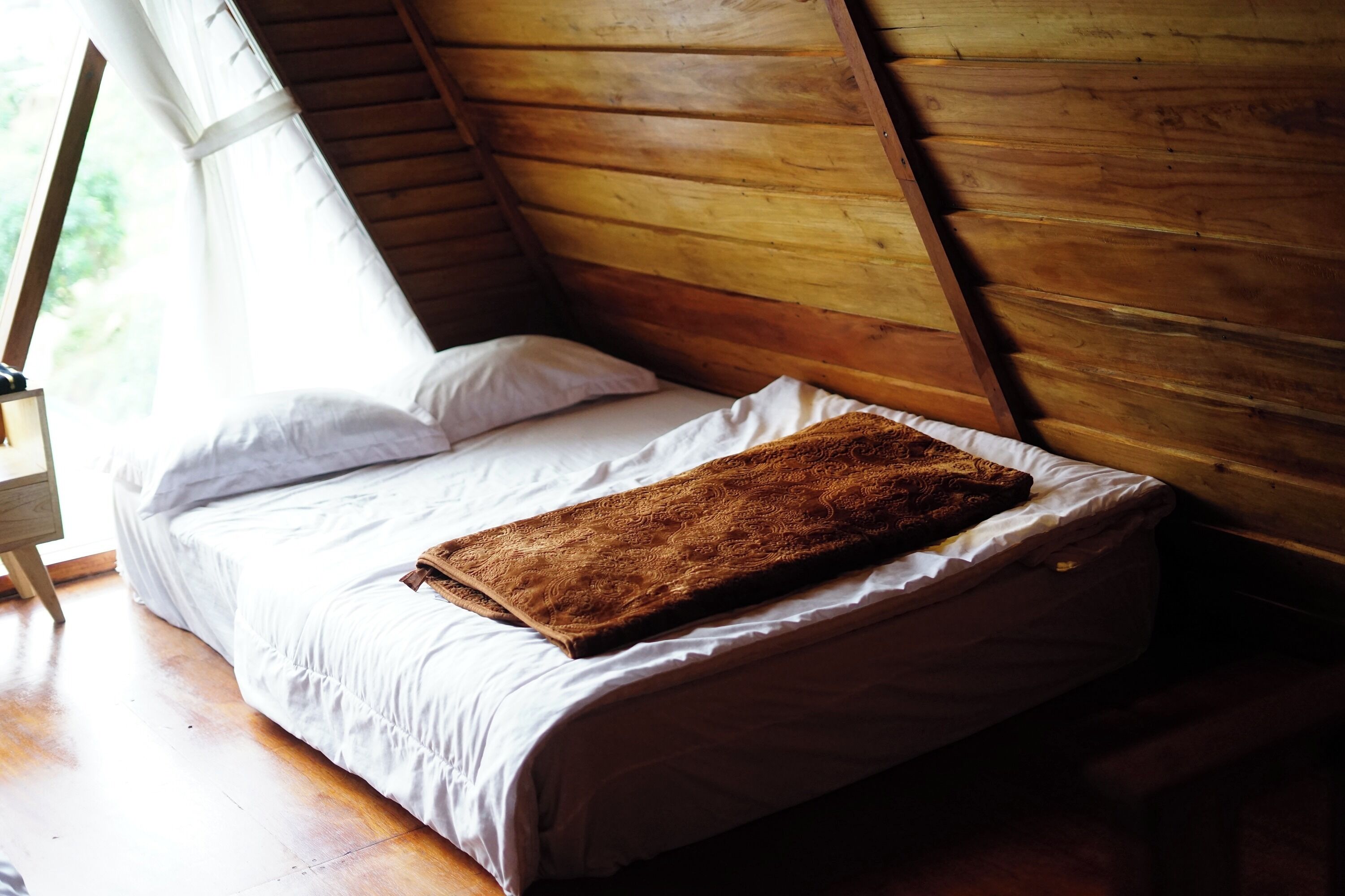 Uma cama de solteiro com roupa de cama branca e um cobertor marrom num quarto com paredes de madeira e uma janela.