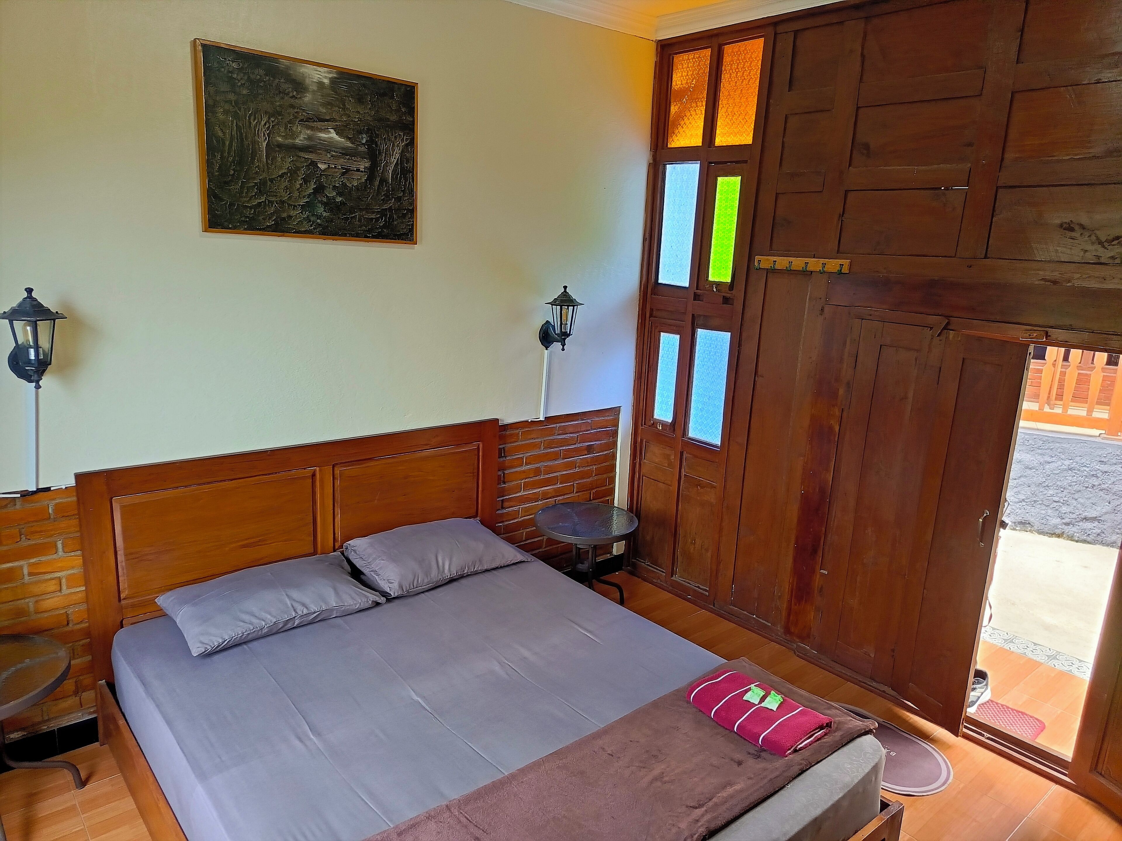 Un dormitorio con cabecera de madera, cama con ropa de cama gris, una mesita, una banca, una lámpara de pared y una puerta de madera con ventanas.