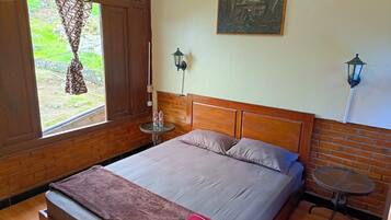 A bedroom with a bed, a small table, a window with curtains, a wall-mounted lamp, and a painting.