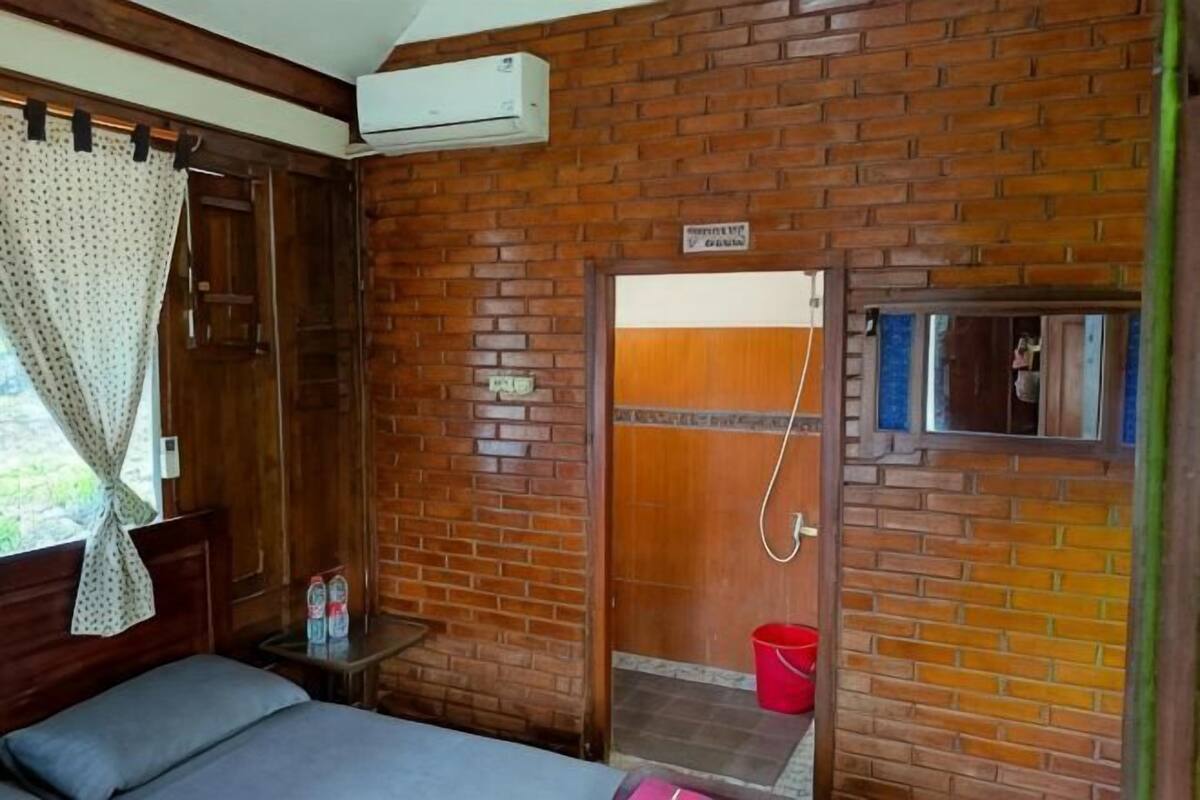 A room with a brick wall, a bed, a small table with a bottle, and a shower area.