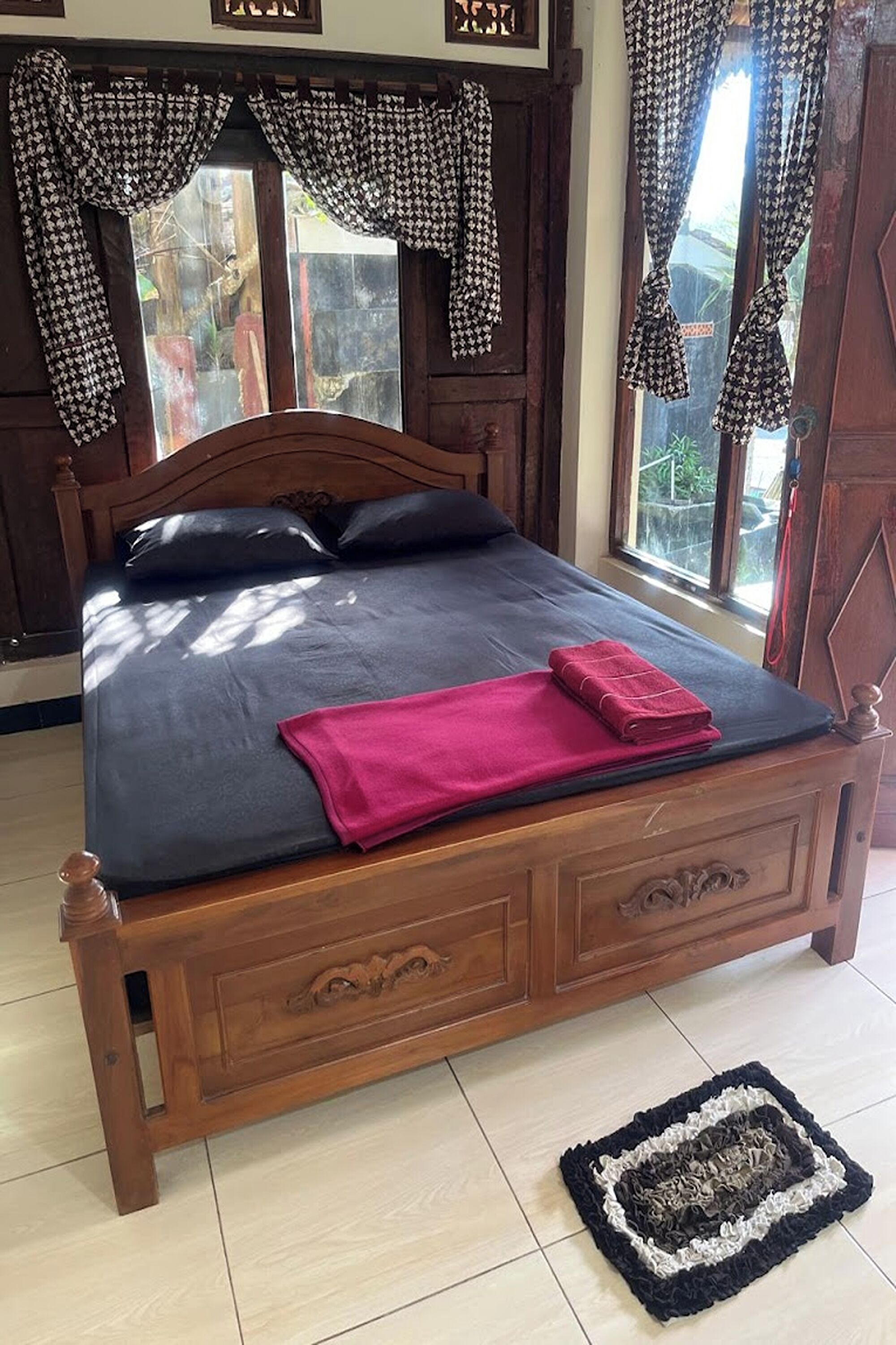A wooden bed with a dark mattress and pillows, a pink blanket, and a black and white rug on the floor.