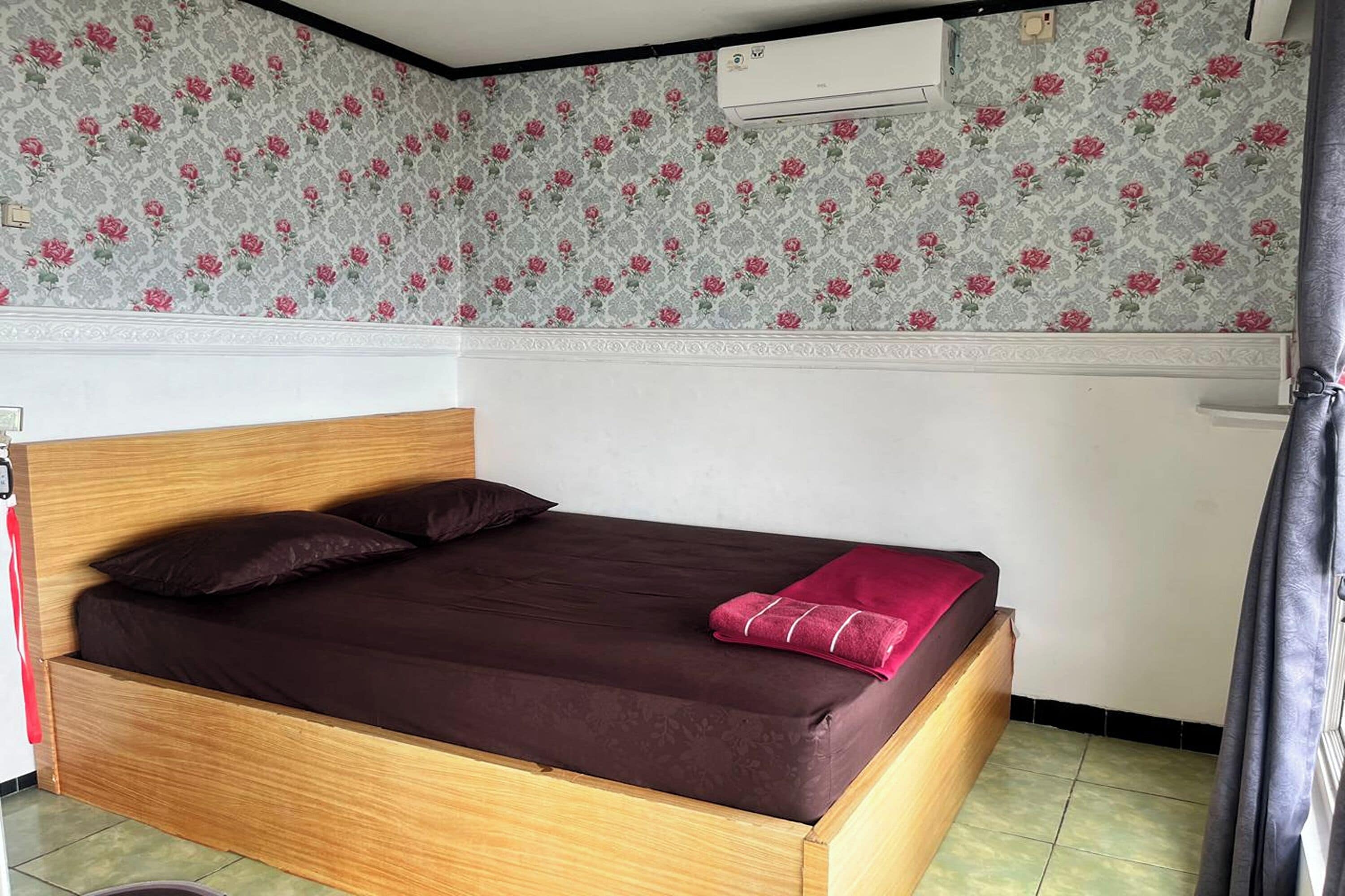 A single bed with a wooden frame and a brown mattress, a red blanket, and a floral-patterned wallpaper.