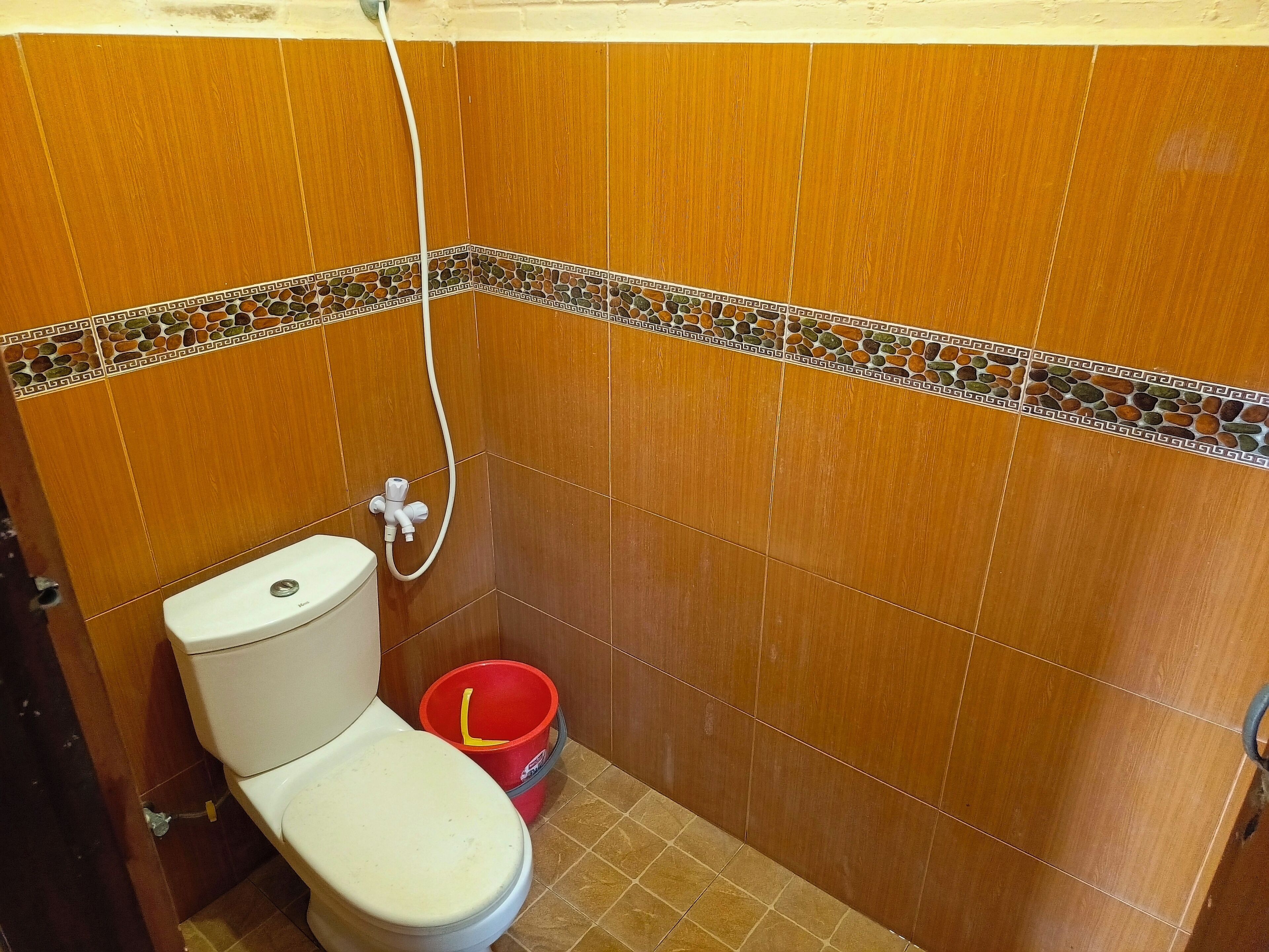 A bathroom with a toilet, a shower hose, and a red bucket.