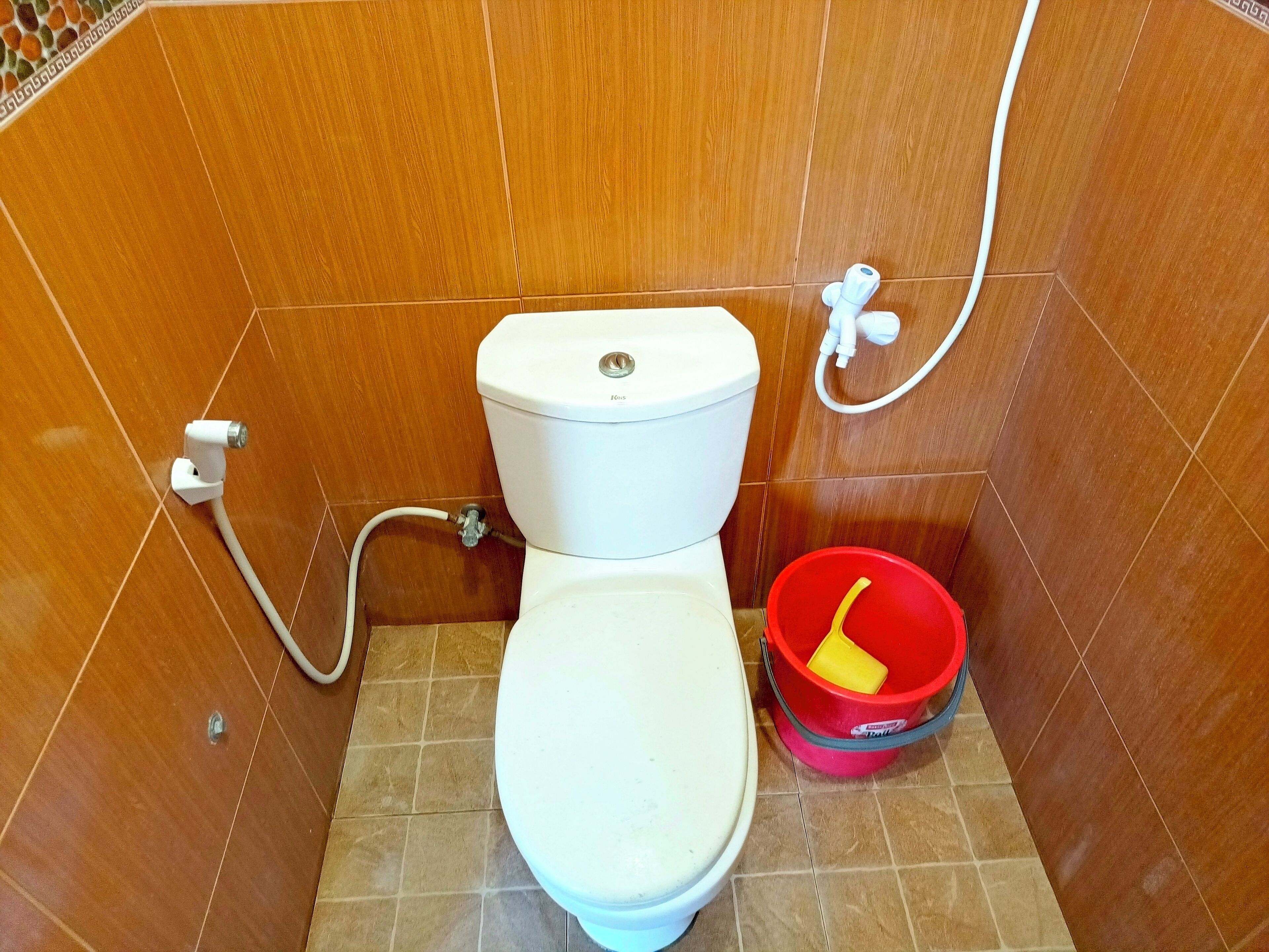 A bathroom with a toilet, a red bucket, and a white pipe.