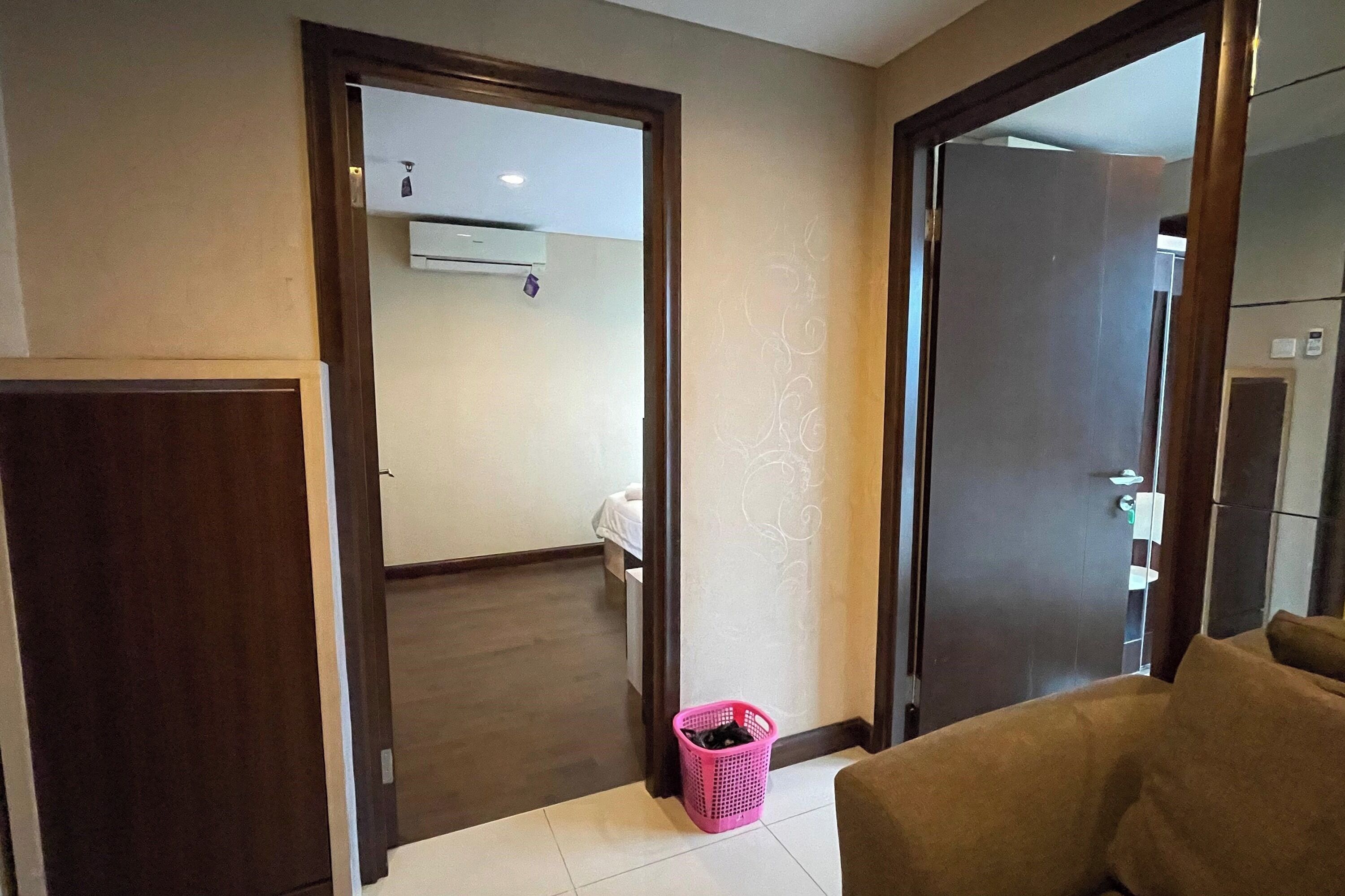 A hotel room with a wooden door, a pink laundry basket, a bed, and a bathroom visible through an open door.