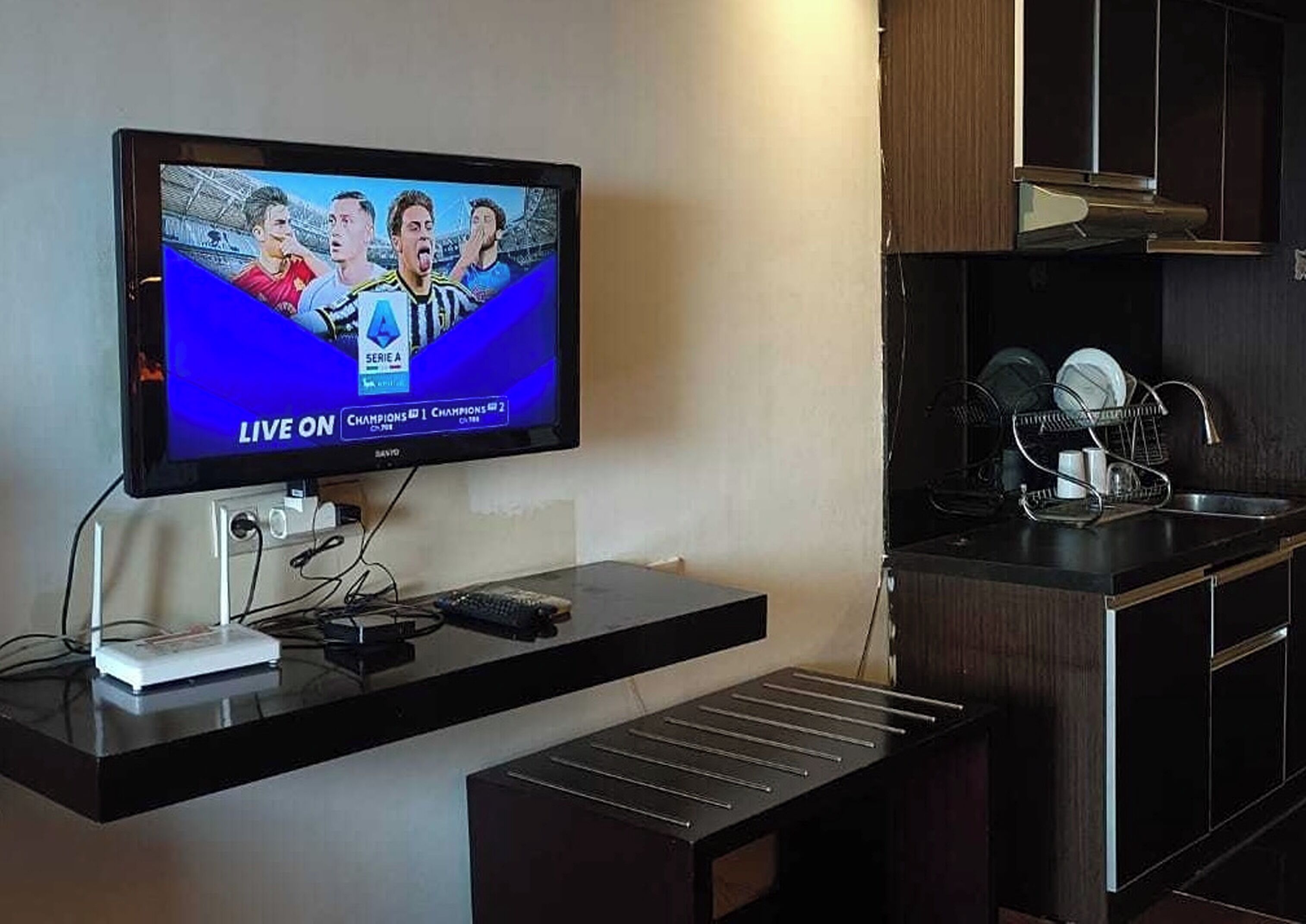 A flat-screen TV mounted on a wall with a live broadcast of a sports event, a black countertop below, and a kitchen area with a sink and dish rack.