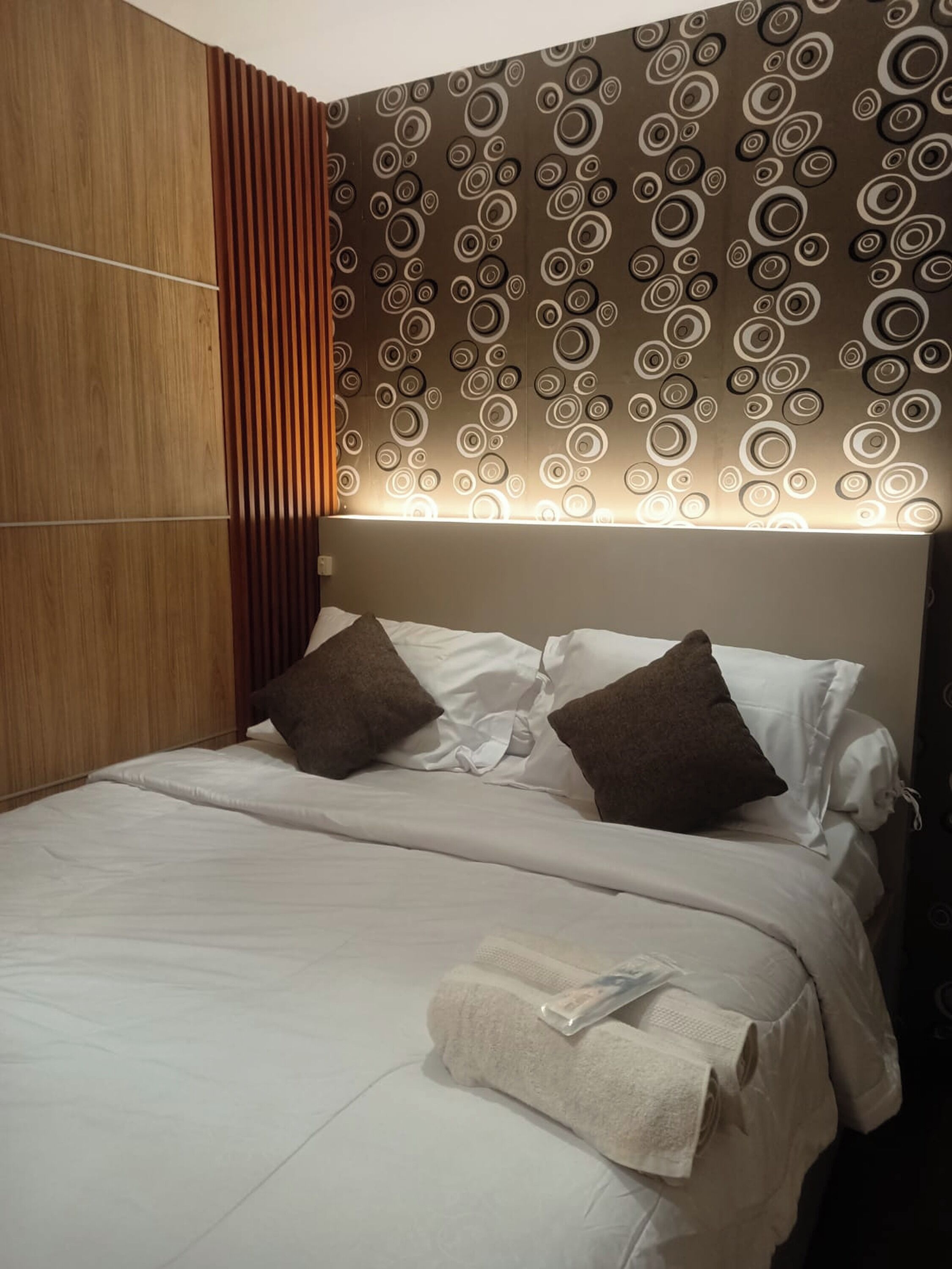A modern bedroom with a wooden panel wall, a circular patterned headboard, and a neatly made bed with pillows and a rolled towel.