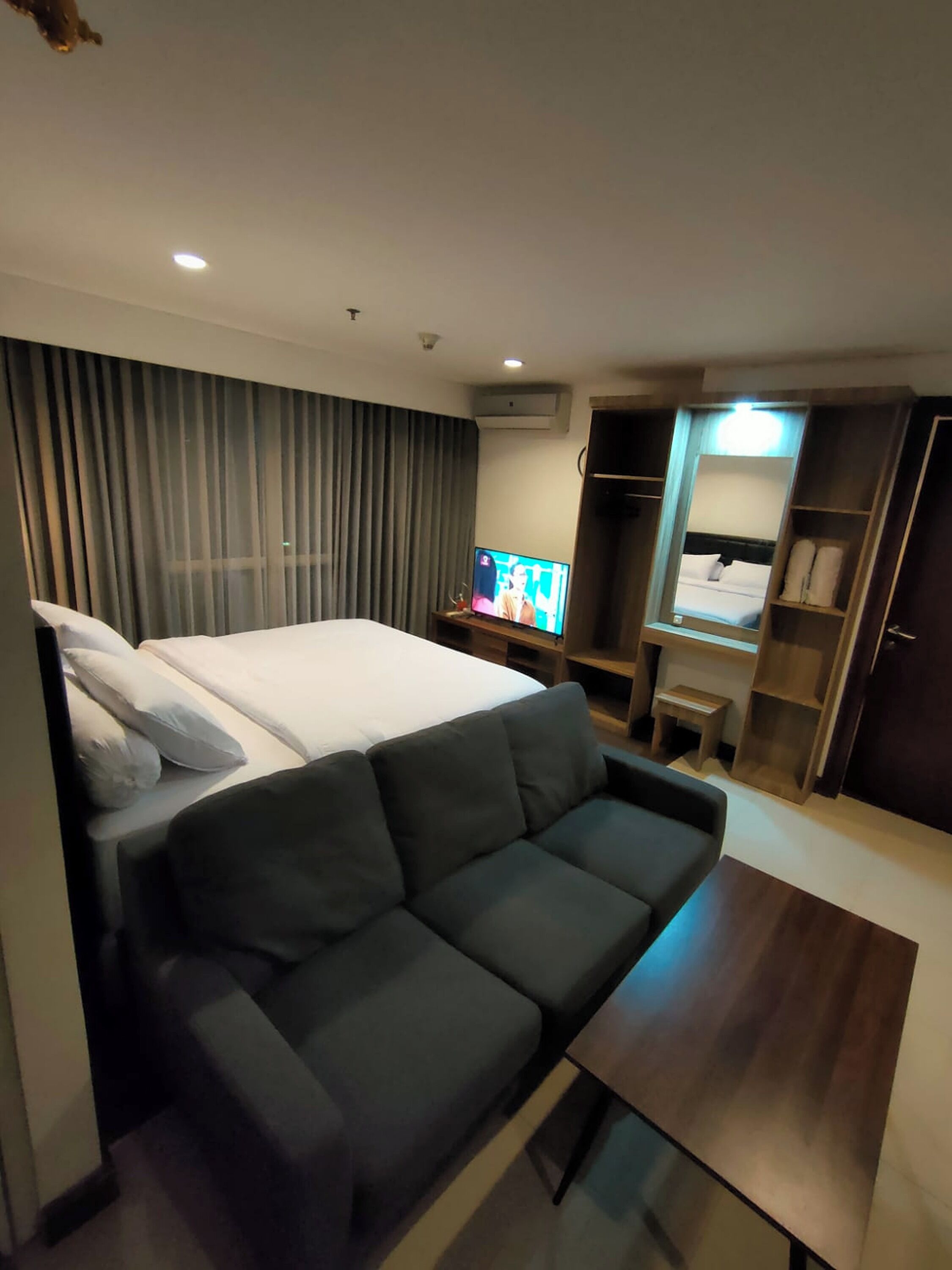 Quarto de hotel moderno com cama, sofá, televisão, escrivaninha e guarda-roupa.