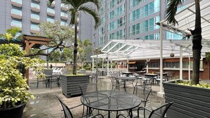 An outdoor patio area with round tables and chairs, surrounded by potted plants and a glass-enclosed structure.