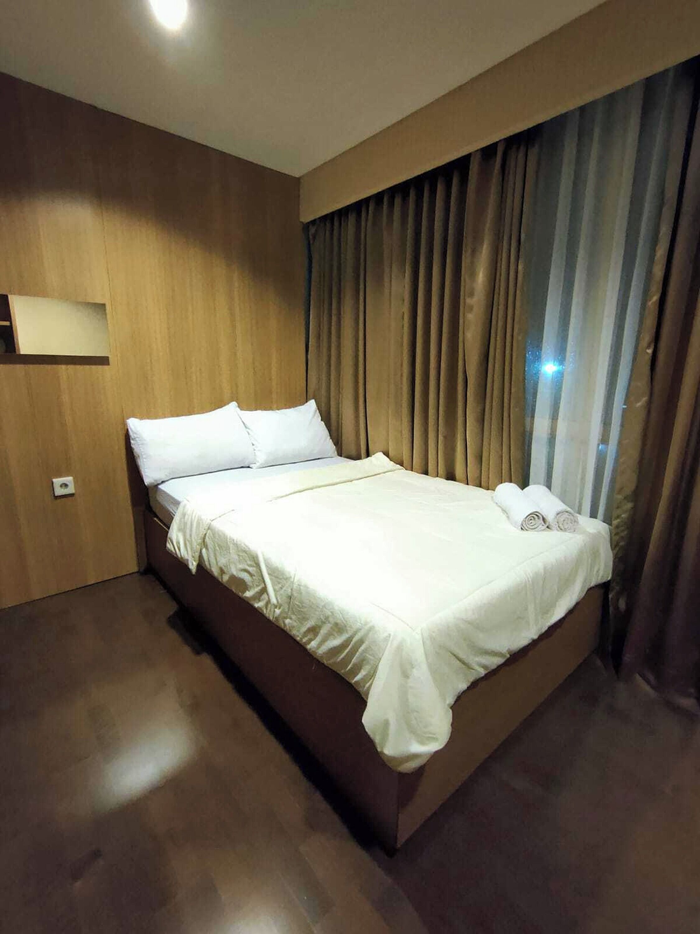 A neatly made bed with white linens and pillows, a wooden headboard, and a large window with curtains.